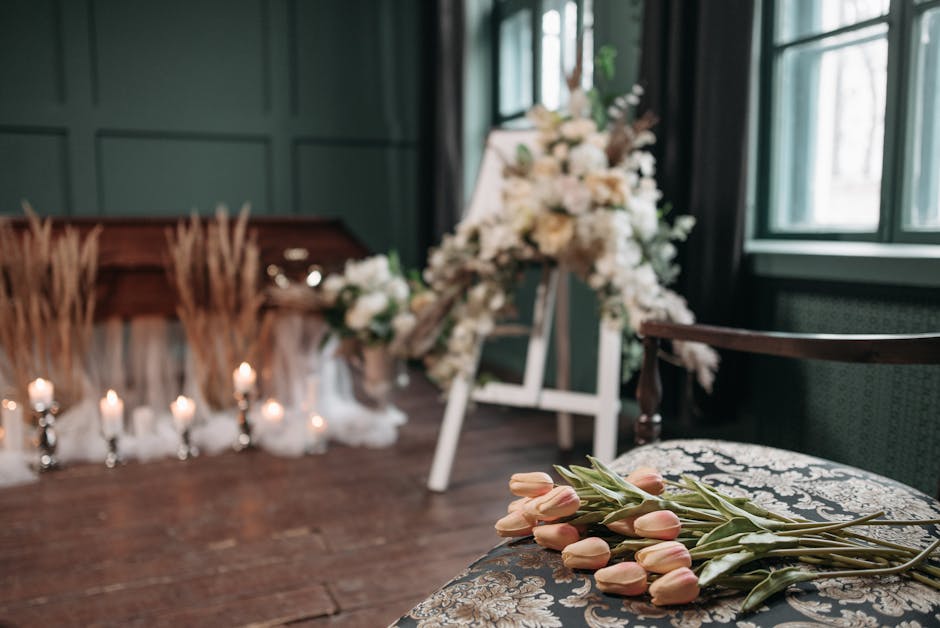 A person dressed in a dark, textured coat is arranging a floral tribute on a polished wooden surface, with a bouquet of pale pink tulips and light green leaves laid out in front. The tulips have smoot