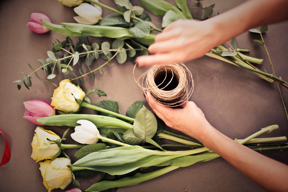A young woman with light skin and long brown hair is arranging a bouquet of fresh flowers in a bright, well-lit flower shop. She is wearing a beige cardigan over a white top and a black skirt with whi