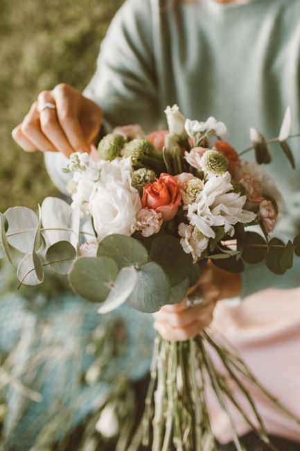 A person in a grey hoodie, with rolled-up sleeves revealing a tattooed arm, is arranging a floral bouquet on a wooden work surface. The bouquet includes white calla lilies, white roses, green foliage,