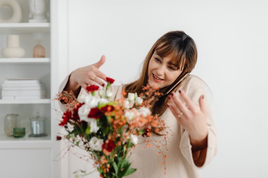 A woman with medium-length brown hair and a cheerful expression, dressed in a light-colored sweater, holding a vibrant bouquet of fresh flowers while speaking on a mobile phone. The bouquet features a