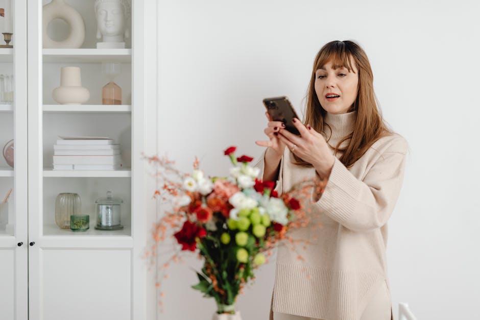 A woman with medium-length brown hair and a cheerful expression, dressed in a light-colored sweater, holding a vibrant bouquet of fresh flowers while speaking on a mobile phone. The bouquet features a