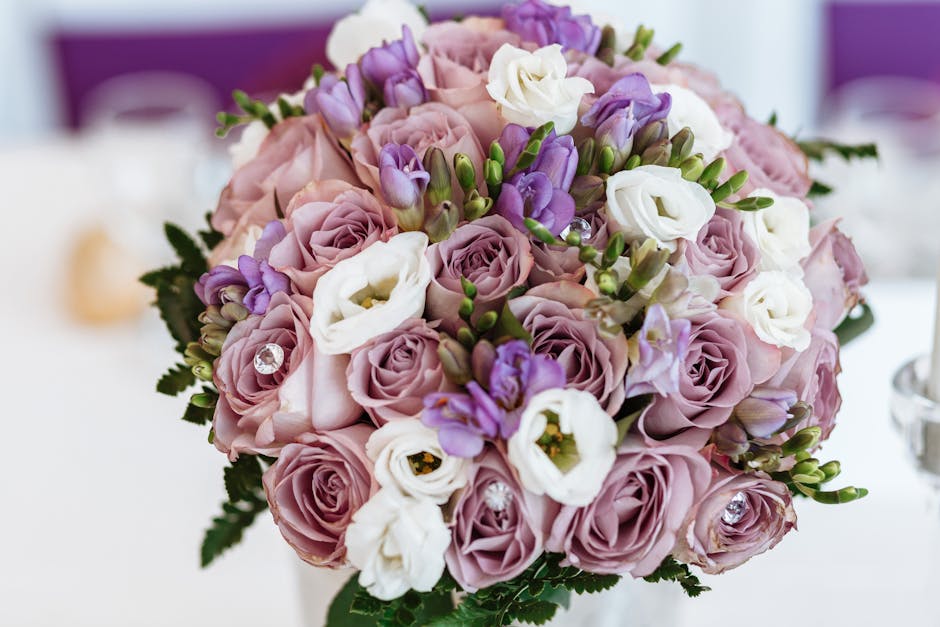 A woman holding a bouquet of freshly arranged flowers, including soft pink roses, white chrysanthemums, and small white filler flowers, wrapped in cream-colored paper with a black label. She is outdoo