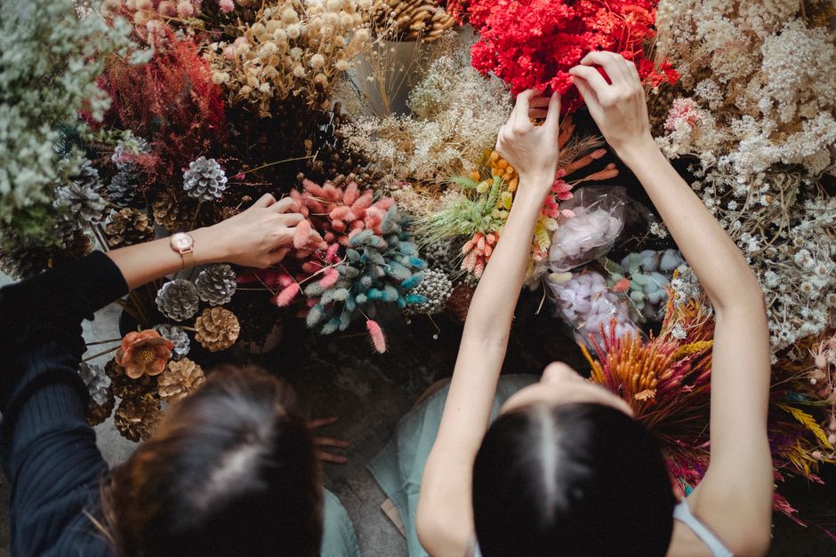 Two women are arranging and inspecting a variety of fresh flowers in a flower shop. The bouquet includes pink roses, white lilies, and other colorful blooms, set in a white ceramic vase. Overhead, bou