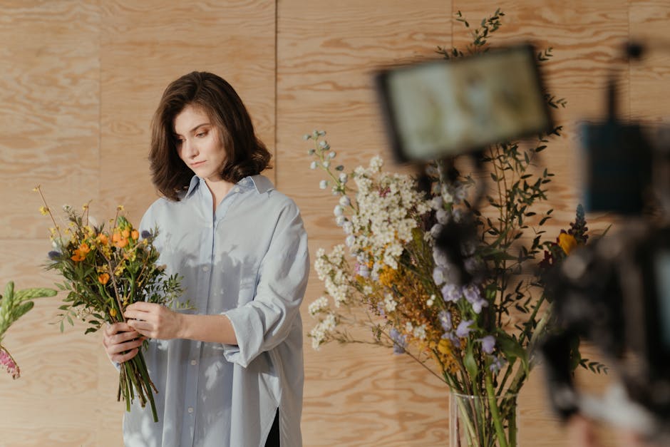 A young female florist with dark hair, dressed in a light gray apron, is arranging pale pink carnations into a white ceramic watering can. She is in a flower shop, surrounded by various floral arrange