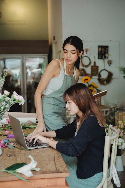 A woman with shoulder-length brown hair, wearing a beige apron and a black cardigan, is smiling while watering a bouquet of pink and white carnations arranged in a white ceramic pitcher. She is standi